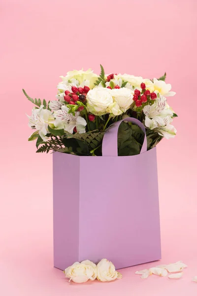 Bouquet of flowers in violet paper bag near petals on pink background — Stock Photo