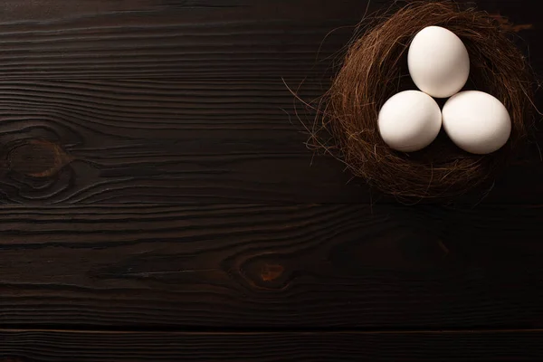 Vue du dessus des œufs de poule blancs dans un nid brun sur fond de bois foncé — Photo de stock