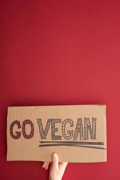 Visão parcial da mulher segurando papelão com go inscrição vegan no fundo vermelho — Fotografia de Stock