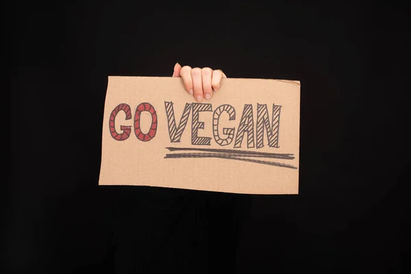 Partial view of woman holding cardboard sign with go vegan inscription isolated on black — Stock Photo