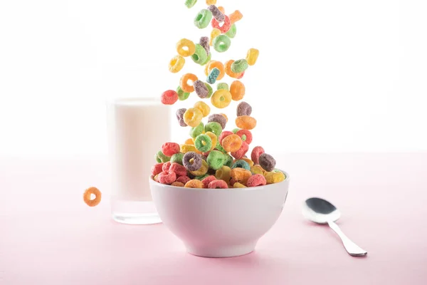 Foyer sélectif de céréales de petit déjeuner multicolores lumineuses tombant dans un bol près du lait et cuillère sur fond blanc — Photo de stock