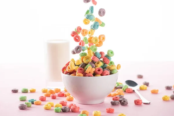 Enfoque selectivo de cereales de desayuno multicolores brillantes que caen en un tazón cerca de la leche y la cuchara sobre fondo blanco - foto de stock