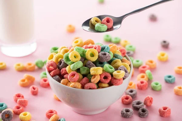 Enfoque selectivo de cereales de desayuno multicolores brillantes en un tazón cerca del vaso de leche y cuchara sobre fondo rosa - foto de stock