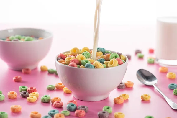 Enfoque selectivo de cereales de desayuno multicolores brillantes en un tazón con leche de verter cerca de la cuchara sobre fondo rosa - foto de stock