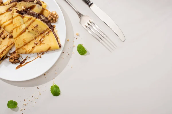 Vue de dessus de crêpes savoureuses avec tartinade au chocolat et noix sur plaque près de couverts et feuilles de menthe sur fond gris — Photo de stock