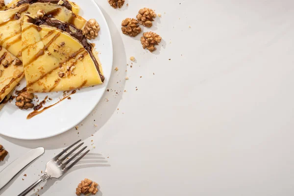 Blick von oben auf leckere Crêpes mit Schokoladenaufstrich und Walnüssen auf Teller neben Besteck auf grauem Hintergrund — Stockfoto