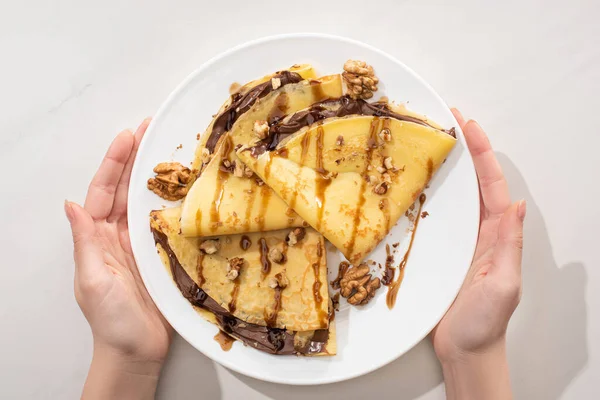 Vista ritagliata di donna con crepes gustose con diffusione di cioccolato e noci su piatto su sfondo grigio — Foto stock