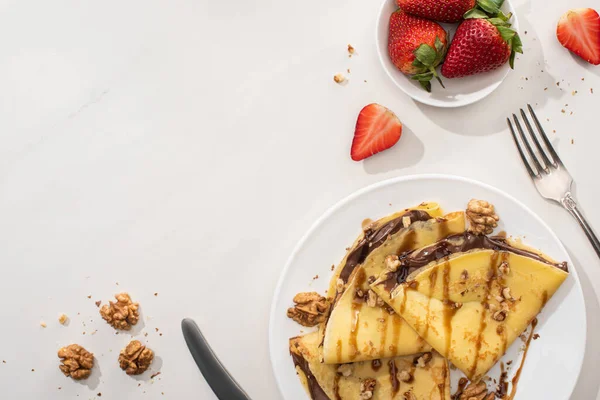 Blick von oben auf leckere Crêpes mit Schokoladenaufstrich und Walnüssen auf Teller neben Schüssel mit Erdbeeren und Besteck auf grauem Hintergrund — Stockfoto
