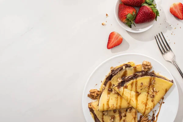 Draufsicht auf leckere Crêpes mit Schokoladenaufstrich und Walnüssen auf Teller neben Schüssel mit Erdbeeren und Gabel auf grauem Hintergrund — Stockfoto