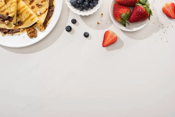 Top view of tasty crepes with chocolate spread and walnuts on plate near bowls with blueberries and strawberries on grey background — Stock Photo