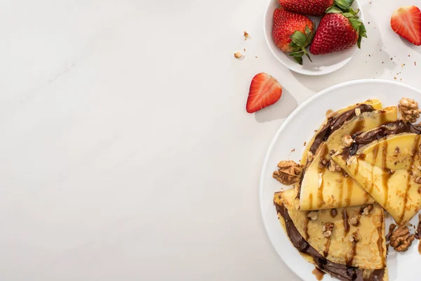 Вид сверху на вкусные блины с шоколадом и грецкими орехами на тарелке возле чаши с клубникой на сером фоне — стоковое фото