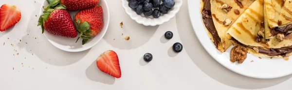 Vue de dessus des crêpes savoureuses avec tartinade au chocolat et noix sur assiette près des bols avec des bleuets et des fraises sur fond gris, vue panoramique — Photo de stock
