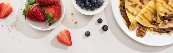 Vista dall'alto di gustose crepes con crema al cioccolato e noci sul piatto vicino a ciotole con mirtilli e fragole su fondo grigio, colpo panoramico — Foto stock