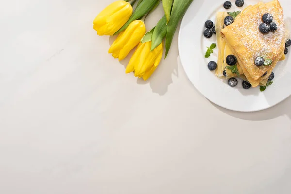 Vista superior de crepes saborosos com mirtilos e hortelã na placa perto de tulipas amarelas no fundo cinza — Fotografia de Stock
