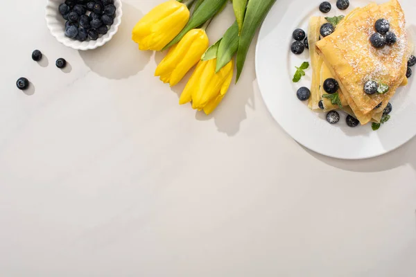 Vista superior de crepes saborosos perto de tigela de mirtilos e tulipas amarelas no fundo cinza — Fotografia de Stock