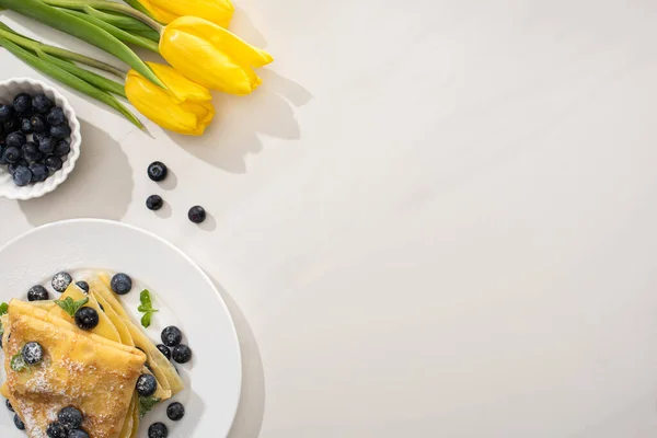 Blick von oben auf leckere Crêpes mit Blaubeeren und Minze auf Teller neben gelben Tulpen auf grauem Hintergrund — Stockfoto