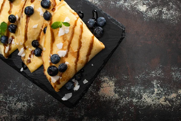 Top view of tasty crepes with blueberries, mint and coconut flakes served on board on textured background — Stock Photo