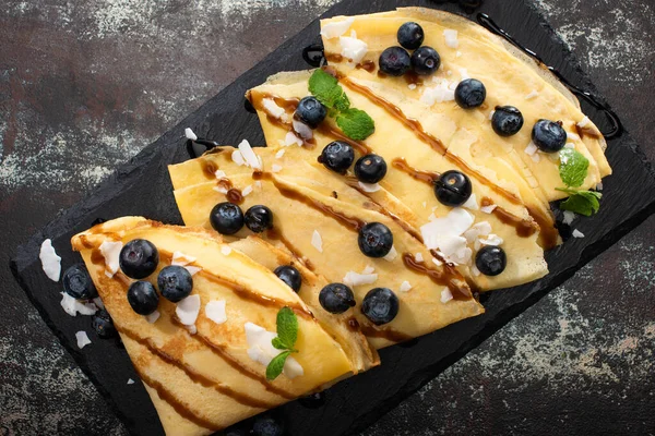 Vue de dessus de crêpes savoureuses aux bleuets, menthe et flocons de noix de coco servis à bord sur fond texturé — Photo de stock