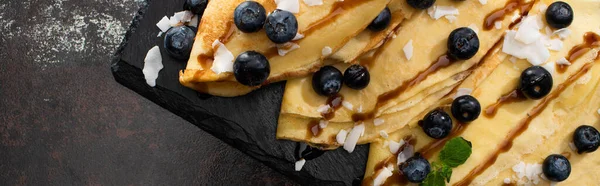 Blick von oben auf leckere Crêpes mit Blaubeeren, Minze und Kokosflocken an Bord auf strukturiertem Hintergrund, Panoramaaufnahme — Stockfoto