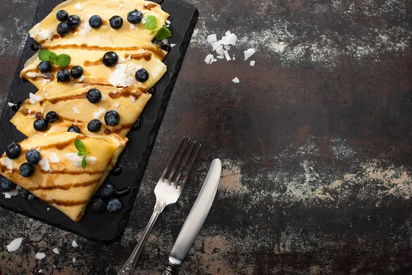 Vista superior de crepes sabrosos con arándanos, menta y copos de coco servidos a bordo cerca de cubiertos sobre fondo texturizado - foto de stock