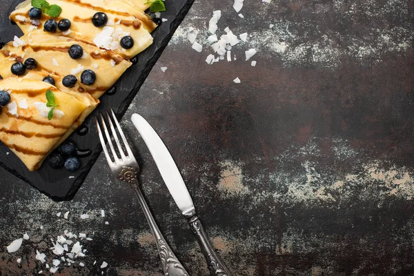 Vista dall'alto di gustose crepes con mirtilli, menta e scaglie di cocco servite a bordo vicino a posate su sfondo strutturato — Foto stock