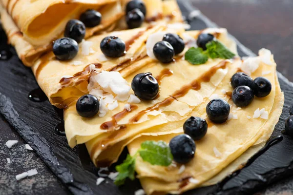 Close up view of tasty crepes with blueberries, mint and coconut flakes served on board on textured background — Stock Photo