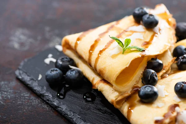 Vista da vicino di gustose crepes con mirtilli, menta e scaglie di cocco servite a bordo su sfondo strutturato — Foto stock