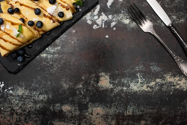 Vista dall'alto di gustose crepes con mirtilli, menta e scaglie di cocco servite a bordo vicino a posate su sfondo strutturato — Foto stock