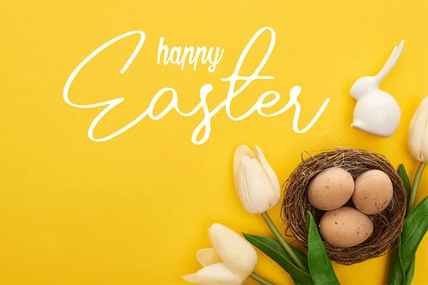 Top view of tulips and chicken eggs in nest near Easter bunny on colorful yellow background with happy Easter illustration — Stock Photo