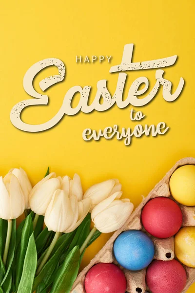 Vue du dessus des tulipes et des oeufs de Pâques peints multicolores dans un récipient en carton sur fond jaune coloré avec une illustration joyeuse de Pâques à tous — Photo de stock