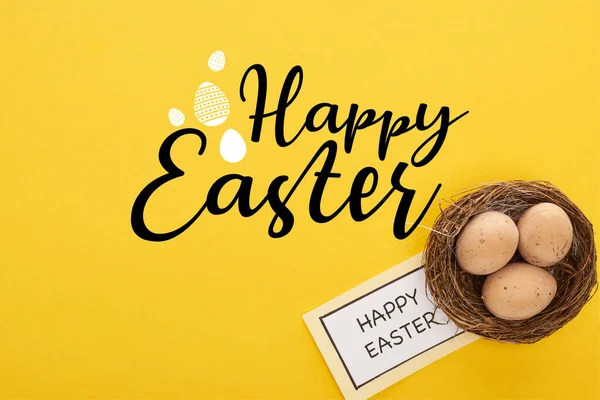 Top view of greeting card with happy Easter lettering near chicken eggs in nest on yellow colorful background — Stock Photo