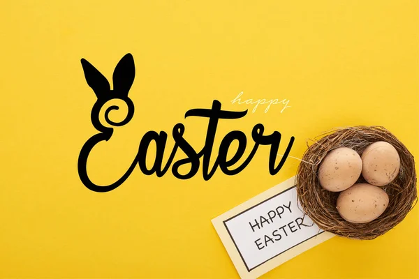 Vista superior de la tarjeta de felicitación con letras de Pascua feliz cerca de huevos de pollo en el nido sobre fondo amarillo colorido - foto de stock