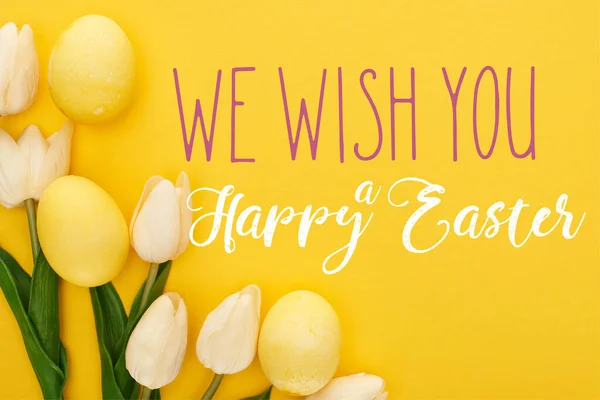 Top view of tulips and painted Easter eggs on colorful yellow background with we wish you a happy Easter illustration — Stock Photo