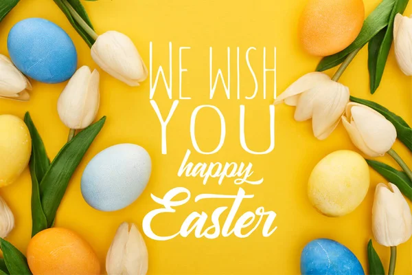 Top view of tulips and painted Easter eggs on colorful yellow background with we wish you a happy Easter illustration — Stock Photo