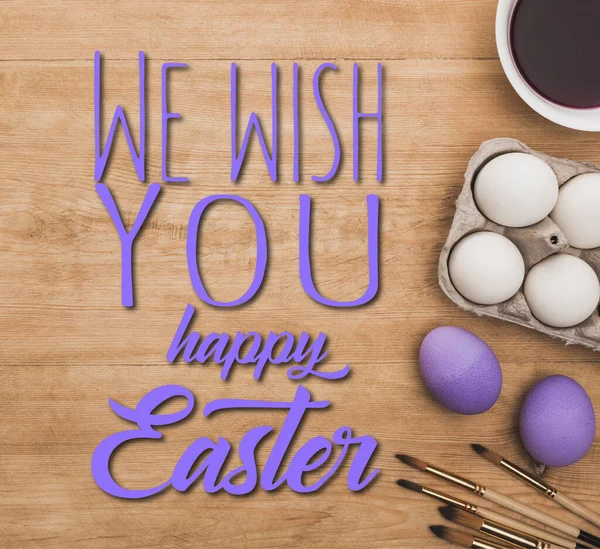 Top view of watercolor purple paint in bowl near chicken eggs and paintbrushes on wooden table with we wish you a happy Easter illustration — Stock Photo
