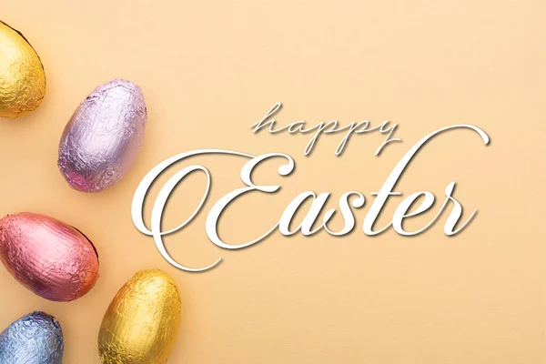 Top view of chocolate Easter eggs in colorful foil on beige background with happy Easter illustration — Stock Photo