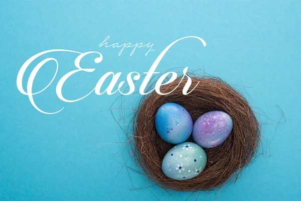 Vista superior del nido con huevos de Pascua púrpura sobre fondo azul con feliz ilustración de Pascua - foto de stock