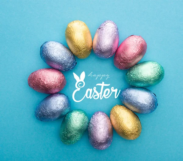 Top view of circle with chocolate Easter eggs in colorful foil on blue background with happy Easter illustration — Stock Photo