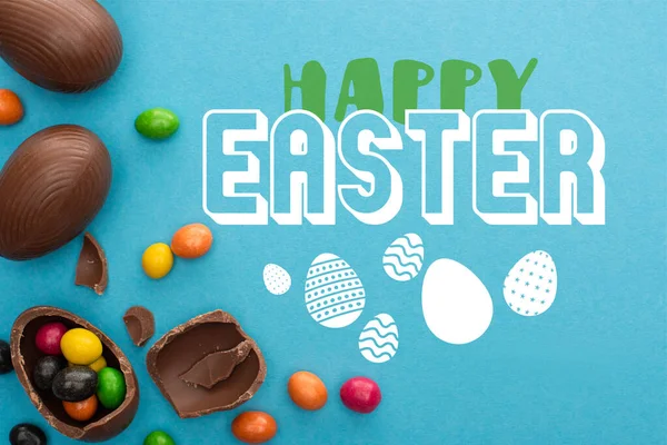 Top view of chocolate Easter eggs with colorful sweets on blue background with happy Easter illustration — Stock Photo