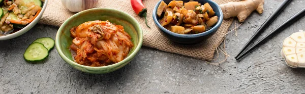 Tiro panorâmico de tigelas com kimchi perto de pauzinhos, gengibre, pimenta, pepino fatiado e alho na superfície de concreto — Fotografia de Stock