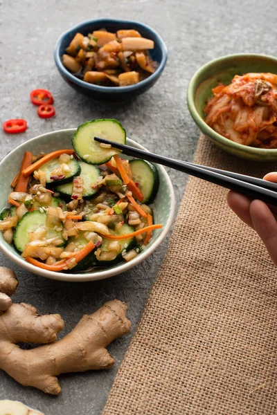 Visão parcial de mulher segurando pauzinhos com pepino fatiado perto de tigelas de kimchi e gengibre na superfície de concreto — Fotografia de Stock