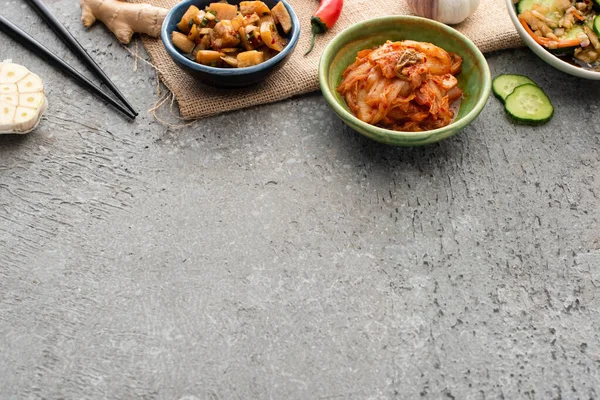 Vista de alto ângulo de tigelas com kimchi perto de pauzinhos, gengibre, pimenta, pepino fatiado e alho na superfície de concreto — Fotografia de Stock