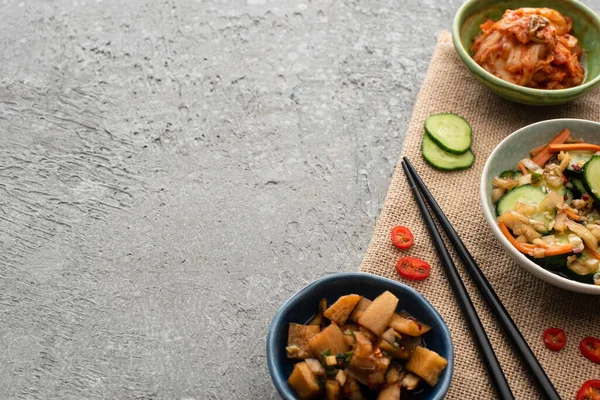 Vista superior de tigelas com kimchi saboroso perto de pauzinhos, pepino fatiado e pimenta em pano de saco na superfície de concreto — Fotografia de Stock