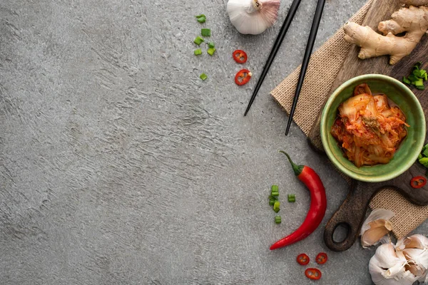 Vista superior da tigela com kimchi na tábua de corte perto de pauzinhos, gengibre, pimenta e alho na superfície de concreto — Fotografia de Stock