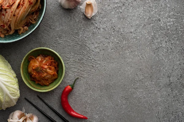 Top view of bowls with kimchi, chopsticks chili pepper, garlic and chinese cabbage on concrete surface — Stock Photo