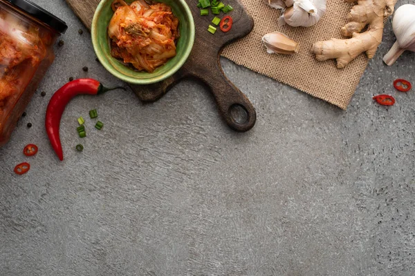 Top view of kimchi in bowl and jar near chili pepper, garlic and ginger on concrete surface — Stock Photo