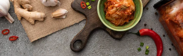 Panoramic shot of kimchi in bowl and jar on cutting board near ginger, garlic and chili pepper on concrete surface — Stock Photo