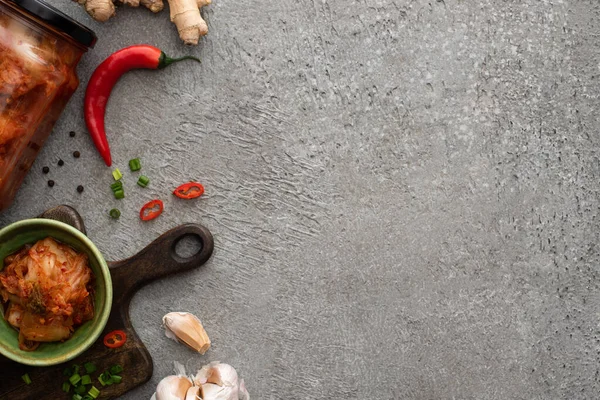 Vue du dessus du kimchi dans un bocal et un bol sur une planche à découper près du gingembre, de l'ail et du piment sur une surface en béton — Photo de stock