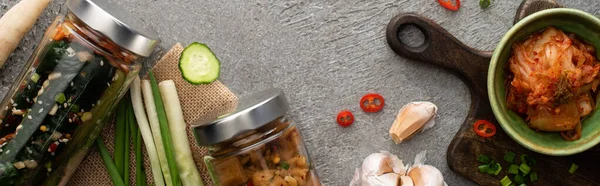 Plano panorámico de sabroso kimchi en frascos y tazón cerca de ajo y cebollas verdes en la superficie de hormigón - foto de stock
