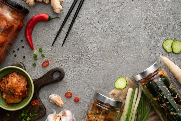 Vue de dessus de savoureux kimchi dans des bocaux et un bol près de baguettes, oignons verts, gingembre, ail et piment sur la surface du béton — Photo de stock